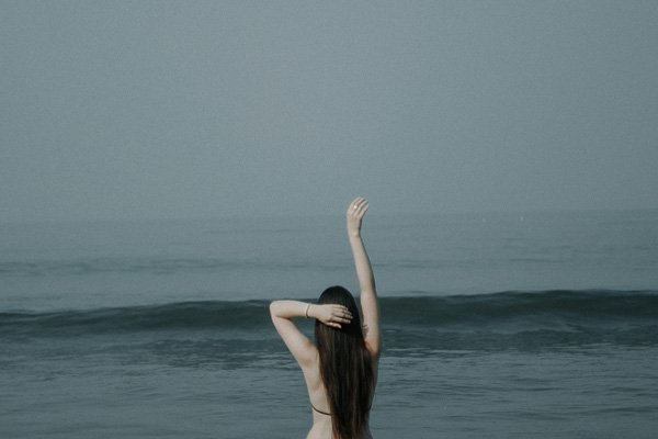 Woman by the sea seen from behind, one arm raised
